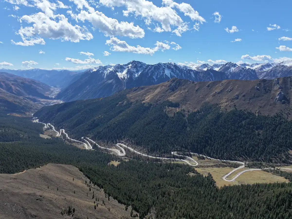 An aerial view of a winding mountain road, dark forest and snow-covered peaks beneath clouds